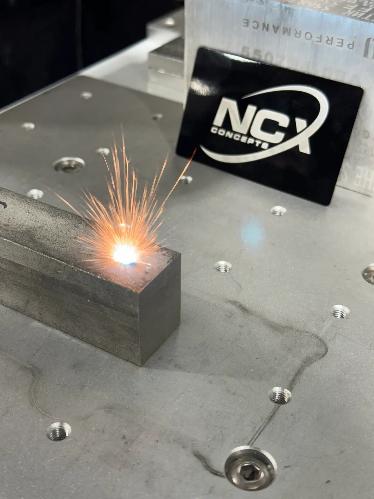 Laser marking a steel block with sparks beside an NCX Concepts logo plate.